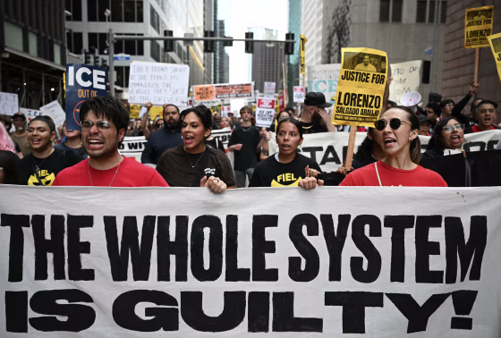 Protestors holding a large white banner reading THE WHOLE SYSTEM IS GUILTY!