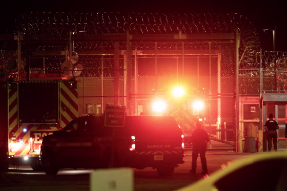 Red lights illuminate the entrance to a prison with tall fencing surrounded by razor wire.