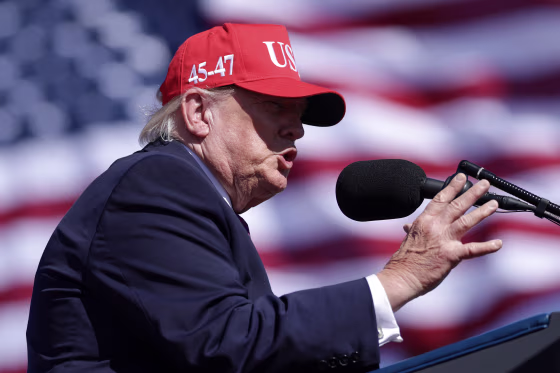 President Trump speaking at a microphone while wearing a red MAGA hat.