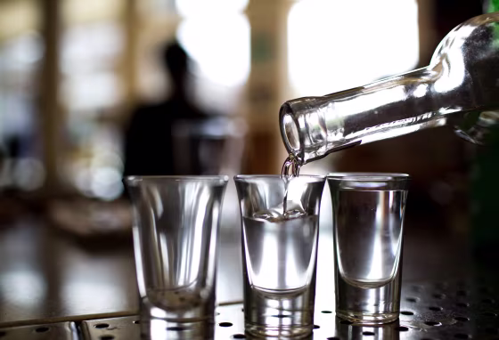 A clear bottle pouring clear liquid into shot glasses against a blurred background.