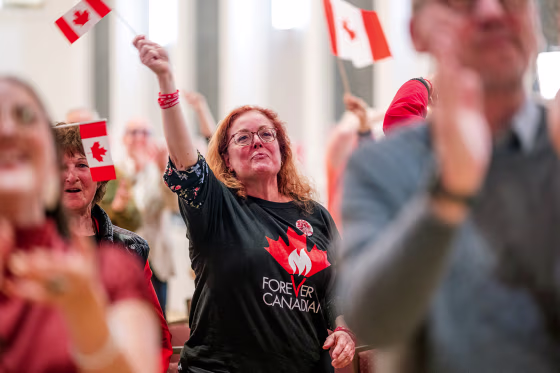 Image: A woman wearing a "Forever Canadian" t-shirt waves a small Canadian flag.