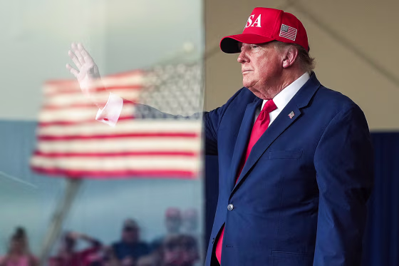 Donald Trump waves, the frame is cut in half by a protective pane of glass that is reflecting an American flag and people in the crowd
