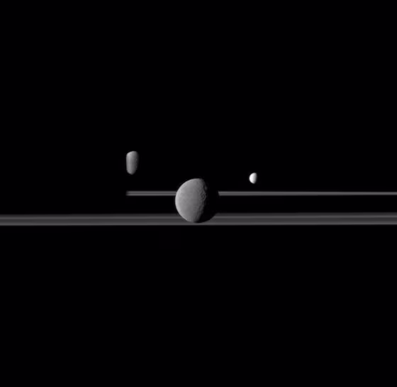 The Cassini spacecraft observes three of Saturn's moons set against the night side of the planet. Saturn is on the left this image but is too dark to see. Rhea (1,528 kilometers, or 949 miles across) is closest to Cassini here and appears largest at the center of the image. Enceladus (504 kilometers, or 313 miles across) is to the right of Rhea. Dione (1,123 kilometers, or 698 miles across) is to the left of Rhea, partly obscured by Saturn.