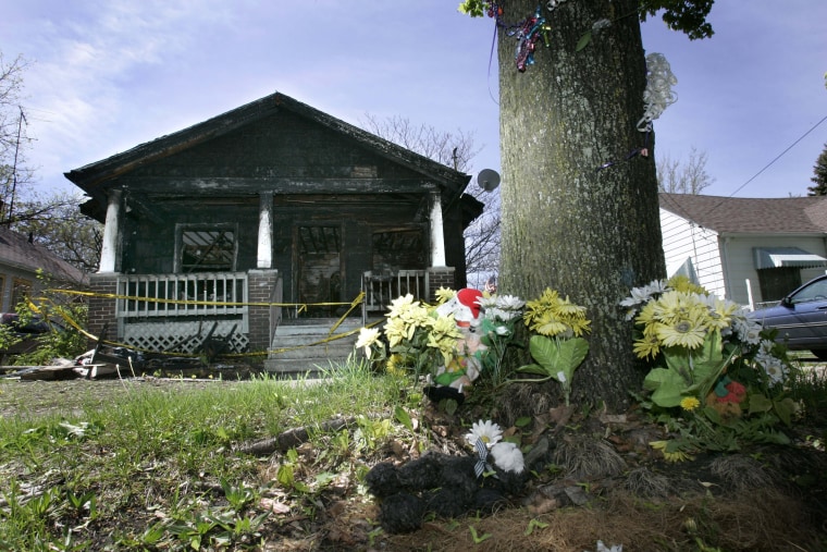 Image: Synthetic flowers at the base of a tree form a temporary memorial to Gordon Yoesting in front of the burned home where he died last October