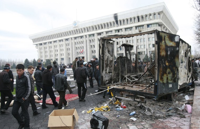 Image: People pass by the burnt remains of a truck in front of \"The White House\", the presidential office, in Bishkek