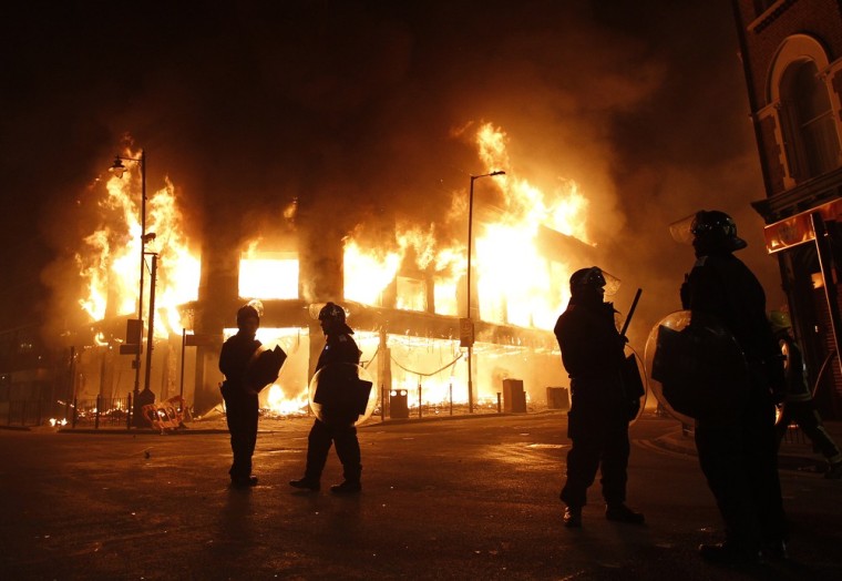 Image: Police officers wearing riot gear stand in front of a burning building in Tottenham