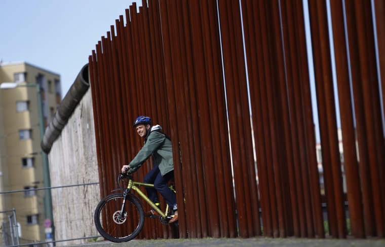 Image: A man rides his bike through gap in row of metal rods that delineates line along which Berlin Wall used to run at Berlin Wall memorial site in Berlin