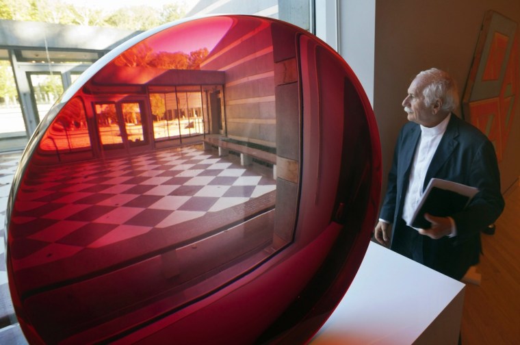 Image: Architect Moshe Safdie looks out of a window next to a large red untitled magnifying disk sculpture by artist Fred Eversley at Crystal Bridges Museum of American Art in Bentonville, Ark.