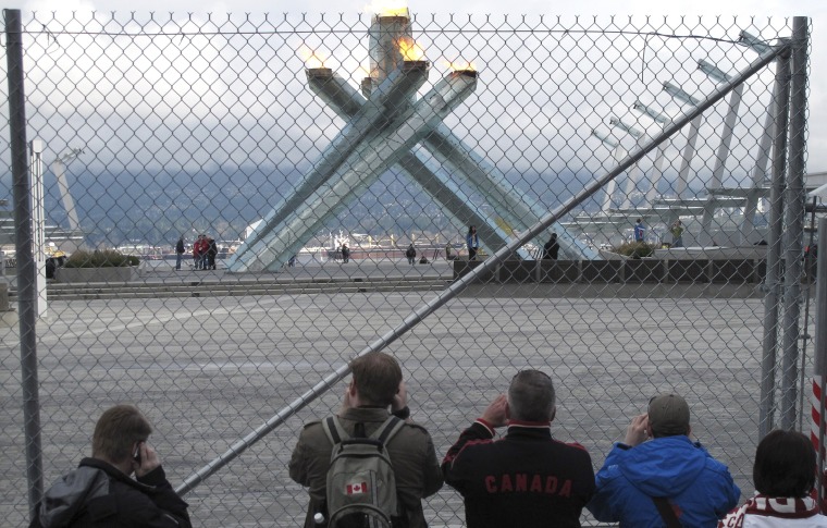 Image: People take pictures of the Olympic cauldron from behind a fence keeping the public out of the plaza it is situated in at the Vancouver 2010 Winter Olympics