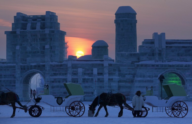 Image: The sun sets behind horses attached to carriages in front of ice sculptures at the 12th Harbin Ice and Snow World display in the northern city of Harbin