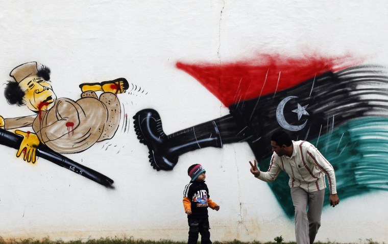 Image: A man plays with his son in front of a cartoon depicting Libyan leader Muammar Gaddafi in Benghazi