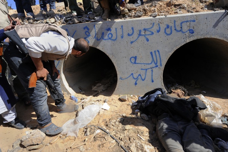 A Libyan National Transitional Council (NTC) fighter looks through a large concrete pipe where Moammar Gadhafi was was captured. A a dead loyalist gunmen lies in the foreground, in Sirte, Oct. 20, 2011.  Arabic graffiti in blue reads: \"This is the place of Gadhafi, the rat.. God is the greatest.\"