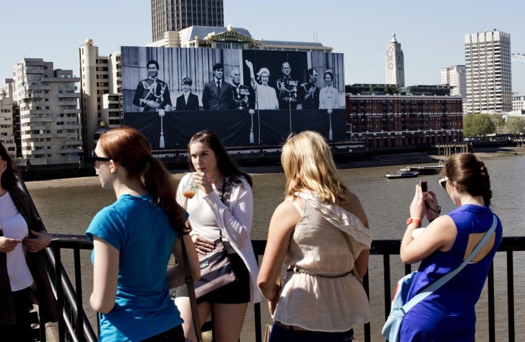 Image: Young women stand across the river as workers help to hang a giant image from a building on the south bank of the River Thames in London showing Britain's Queen Elizabeth II and the royal family.