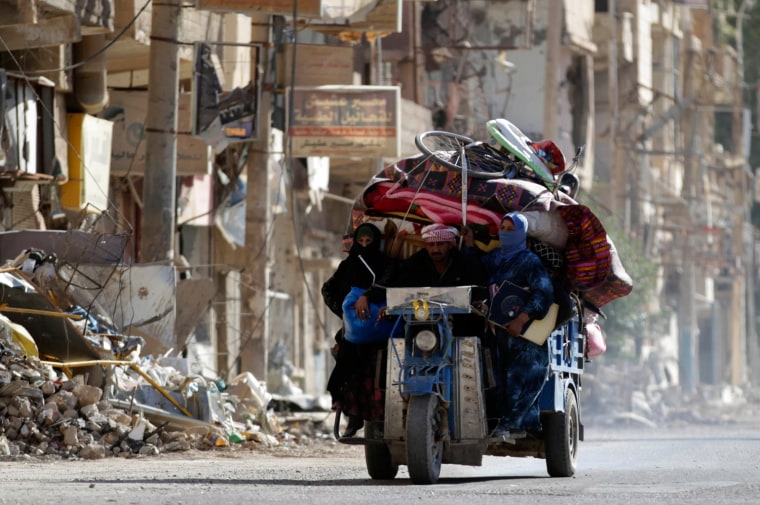 Image: Residents ride on a motorcycle with their belongings past damaged buildings in Deir al-Zor
