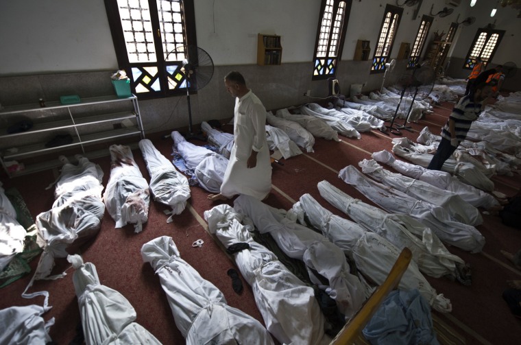 Image: An Egyptian man walks between lines of bodies wrapped in shrouds at a mosque