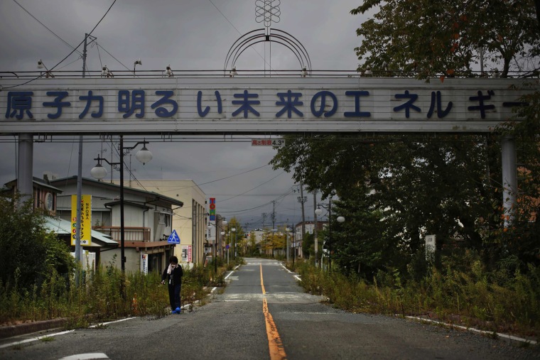 Image: A woman who came for a brief visit to her home walks under a sign reading \"Nuclear Power - The Energy for a Better Future\", at the entrance of empty Futaba town, inside the exclusion zone in Fukushima prefecture