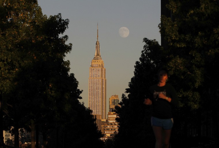 The moon rises at sunset behind New York's Empire State building as a woman runs along a promenade in Hoboken, New Jersey