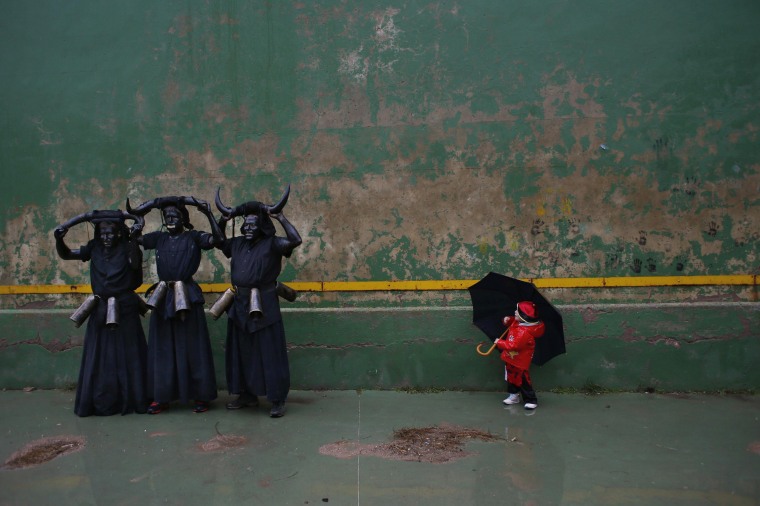Image: A boy looks as men dressed as \"Diablos de Luzon\" (Luzon Devils) pose for a portrait during carnival celebrations in the Spanish village of Luzon