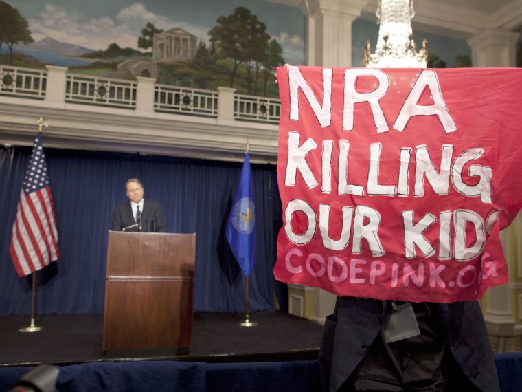 A protester holds up a sign as National Rifle Association Executive Vice President Wayne LaPierre, left, speaks during a news conference in response to the Connecticut school shooting on Friday, Dec. 21, 2012 in Washington. (Photo by Evan Vucci/AP Photo)
