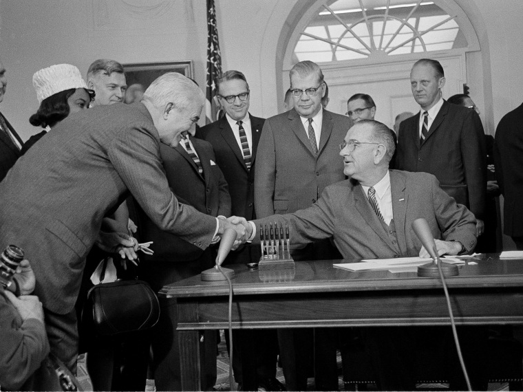 President Lyndon B. Johnson, who used 60 pens to sign a $1.2 billion college aid bill at the White House, hands one of the souvenirs to President Logan Wilson of the American Council on Education, Dec. 16, 1963. A room full of persons who helped pass...