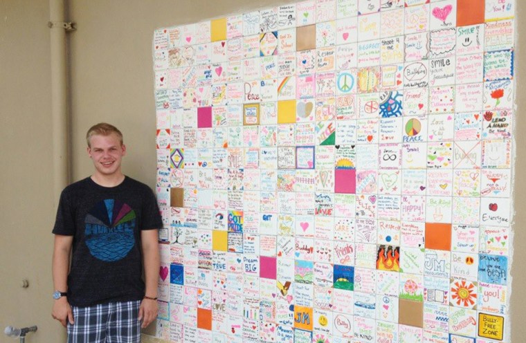 Ryan Andresen stands in front of a \"tolerance wall,\" his final Boy Scouts' project that he worked on with school children at his former middle school. It consists of 288 tiles that depict acts of kindness.