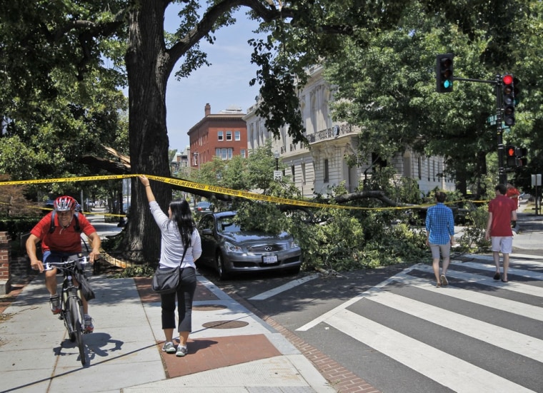 A pedestrian helps a bicyclist navigate a sidewalk blocked by a fallen tree that also damaged a parked vehicle in the Dupont Circle neighborhood of Washington, D.C., Saturday, June 30.