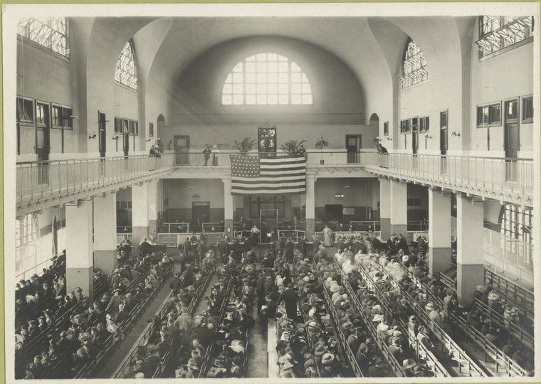 Immigrants seated on long benches, Main Hall, U.S. Immigration Station. The 46-star American flag hanging from the balcony indicates that this photograph was taken between 1907 and 1912.
