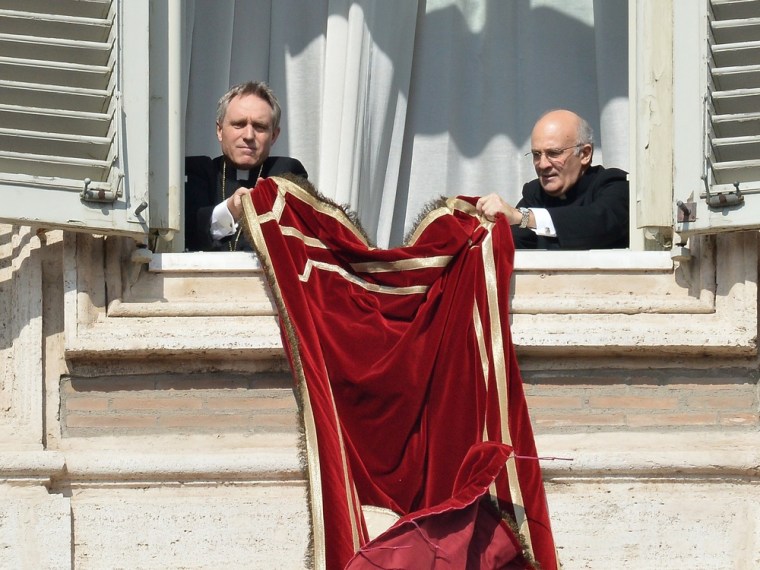 The pope's secretary Georg Gänswein (L) and an unidentified ecclesiastic set the tapestry featuring the tiara on the balcony of St Peter's basilica ahead of Pope Benedict XVI's last Sunday prayers before stepping down on February 24, 2013 at St Peter's...