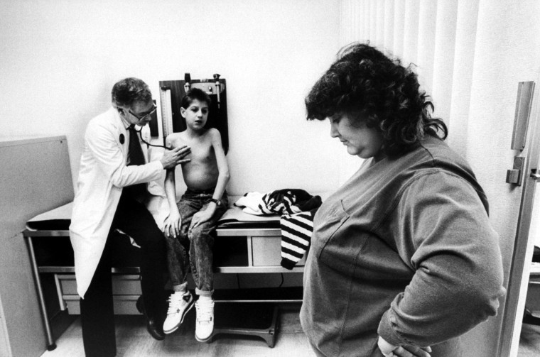 Ryan White sits on an examining table as his physician Dr. Martin Kleiman uses a stethoscope to listen to his lungs, while mom Jeanne looks on at hospital.