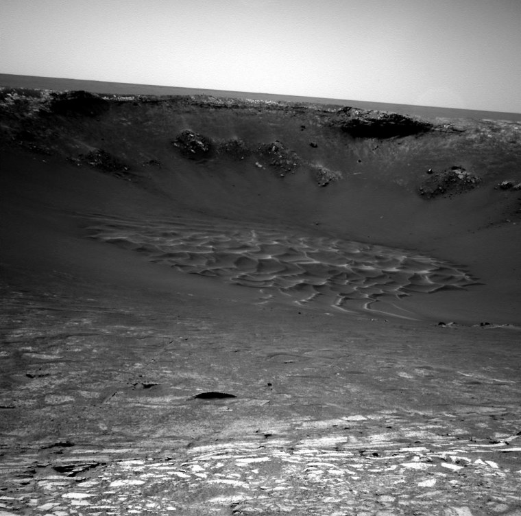 The view from Opportunity's navigation camera shows the downhill slope of Endurance Crater's rim, leading to a sweeping set of sand dunes at the crater's floor. Flat slabs of bedrock are spread out in the foreground.