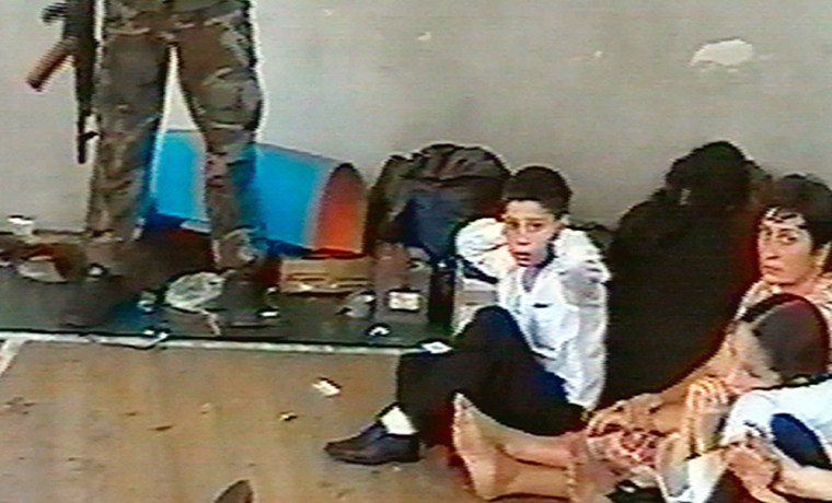 ** RETRANSMISSION OF MOSB801, OF SEPT 1, TO ADD IDENTIFICATION ** Hostages sit on a floor in a gym as a hostage-taker stands with his left foot on a book apparently with a device connected to a chain of explosives in the school in Beslan, taken on an unknown date during the early part of the siege which began on Sept. 1, 2004, in this image from television. Georgy Farniyev is at center, Sima Alikova and her 9-year-old daughter Irina are at right. (AP Photo/NTV-Russian Television Channel) ** TV OUT **