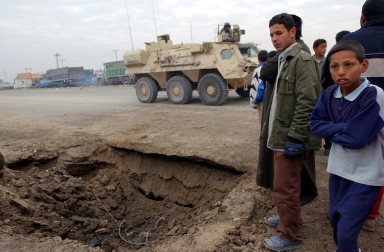 People stand around a crater caused by an explosive device as a US armored personel carrier drives by on Baghdad's outskirts Wednesday Dec. 8, 2004. (AP Photo/Khalid Mohammed)