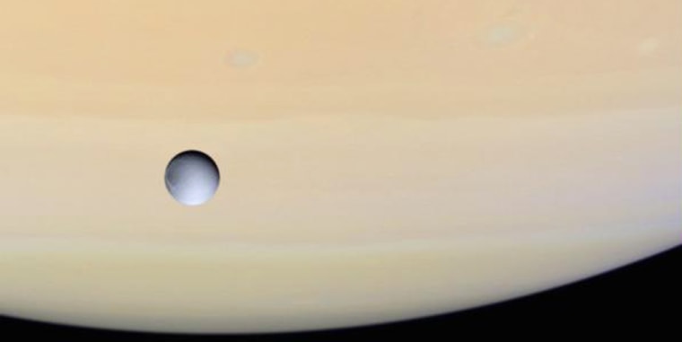 The grayish disk of the moon Dione is set against the pastel clouds of Saturn in this image, captured by the Cassini spacecraft.