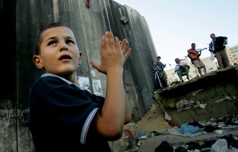 Members of the band Mille Dancer from Italy perform as a Palestinian boy claps as they are backdropped by a section of Israel's separation barrier in the West Bank town of Abu Dis, on the outskirts of Jerusalem, Tuesday July 5, 2005. The band is visiting as part of the Palestinian International Festival that is taking place in Ramallah. (AP Photo/Kevin Frayer)
