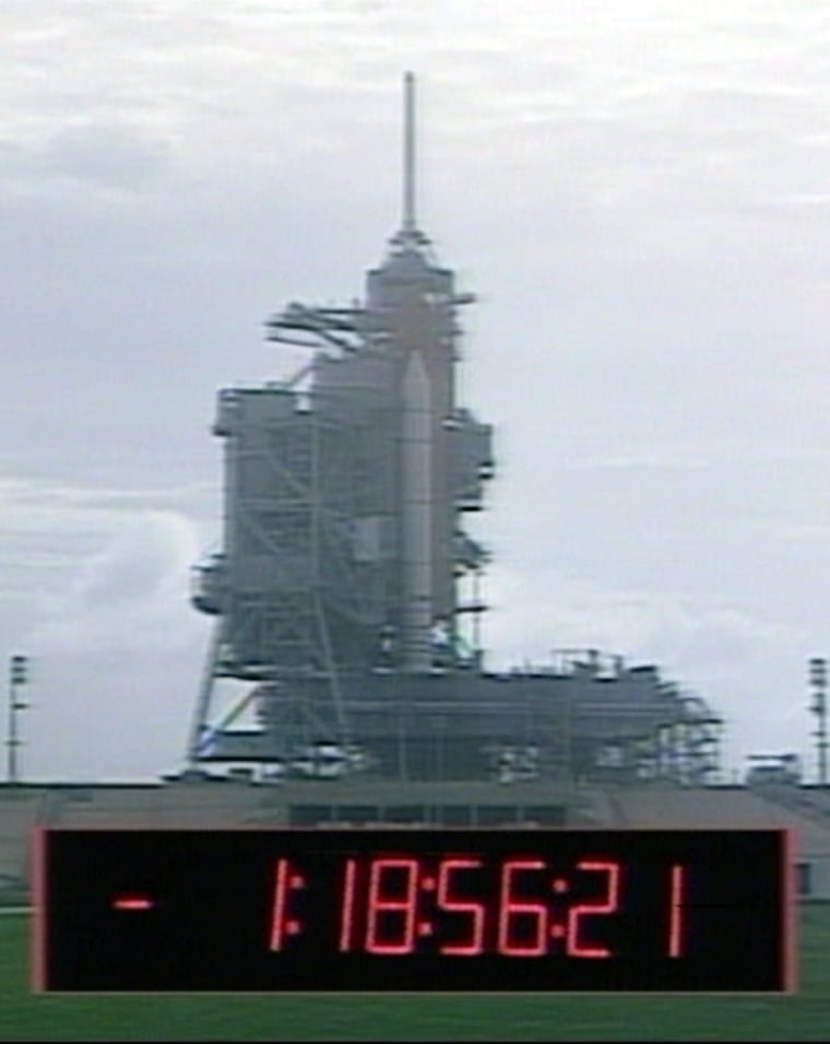 In this image from NASA TV, a virtual countdown clock is superimposed over a view of the shuttle launch pad. The clock shows one day, 18 hours, 56 minutes and 21 seconds. The time is less than the amount of time until the schedule liftoff, because of a series of built-in pauses that halt the countdown.