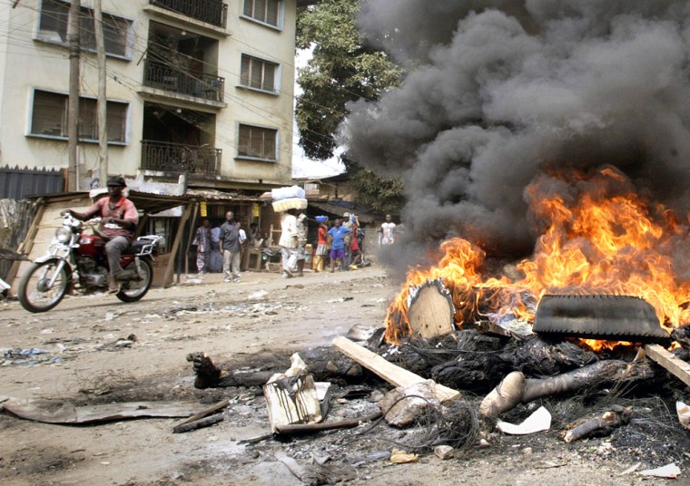 People pass by bodies amid burning tires and debris Thursday in a street in Onitsha, Nigeria. Muslims fled the city and corpses still smoldered in its streets Thursday as two days of sectarian violence that killed scores subsided. 