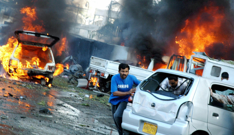 Pakistani people push a car past burning