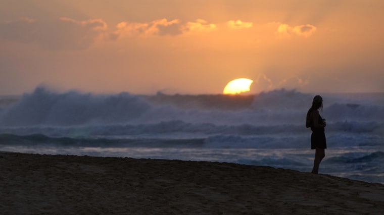 A women watches the sun set behind swellls of waves rolling to the shore at Sunset Beach, Hawaii.