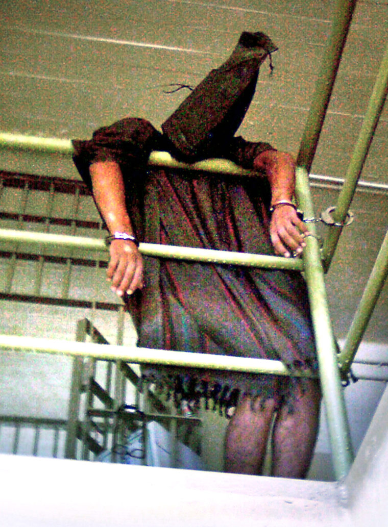 In this undated photo, a hooded Iraqi detainee appears to be cuffed at both wrists and collapsed over a rail at the Abu Ghraib prison.