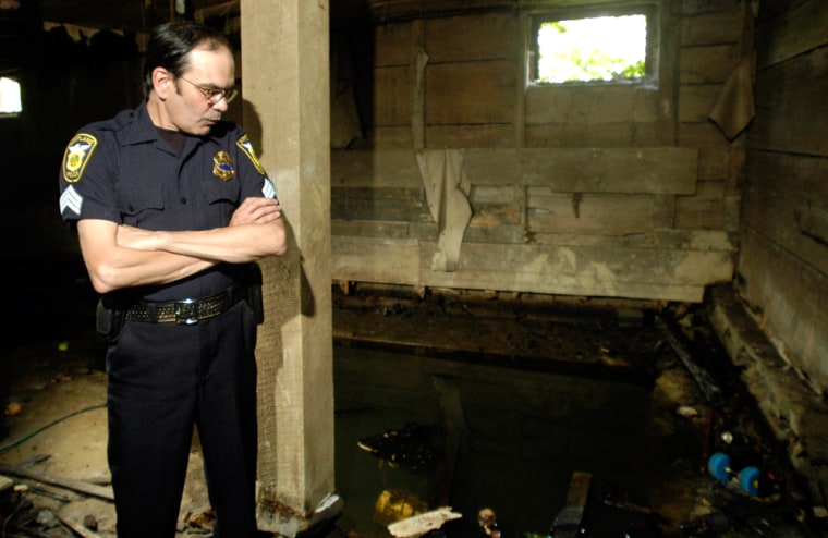 Kirtland police officer Sgt. Ron Andolsek poses for a photo in the barn where the Avery family was murdered by Jeffrey Lundgren in 1989, in what became known as the Kirtland cult slayings, in Kirtland, Ohio, Friday, Aug. 25, 2006. Behind him, now filled with run-off rain water, is the pit where the bodies where buried and Andolsek and other authorities excavated the bodies from. Lundgren's execution is scheduled for Tuesday, Oct. 24, 2006. (AP Photo/Jamie Yanak)