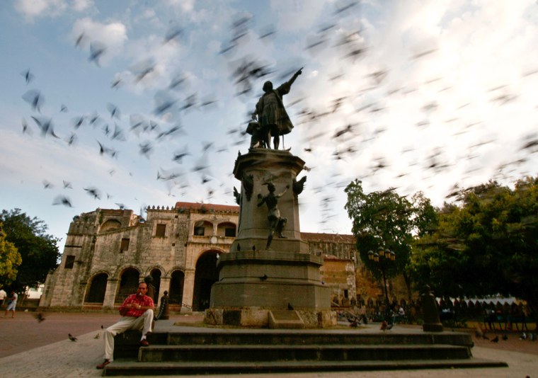 A man sits near a monument as pigeons fly above in Santo Domingo