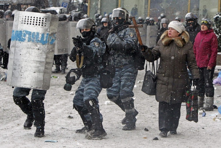 Image: A woman holds a wodden cross as riot policemen aim their weapons during clashes with pro-European protesters in Kiev
