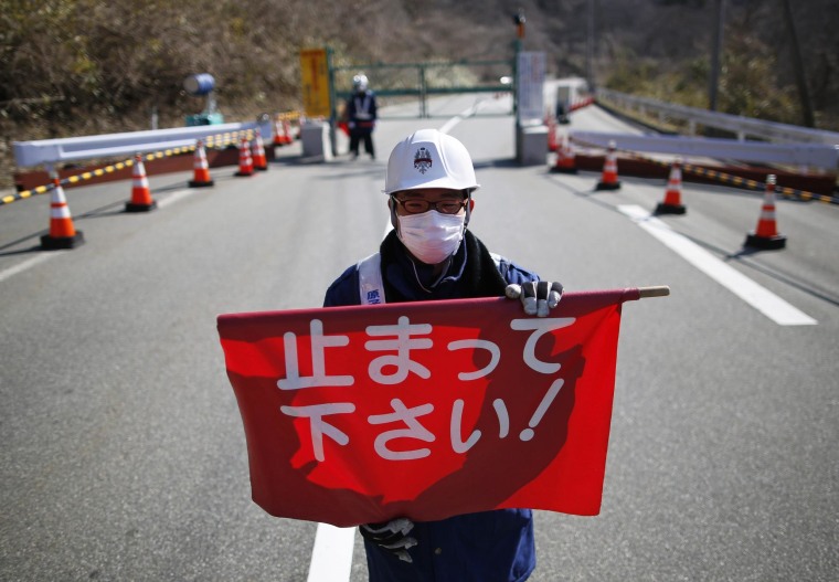 Image: A security personnel holds a flag as he stand by a steel gate that marks the border between Tamura and Okuma town in Okuma town, Fukushima prefecture