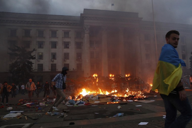 Image: A protester wrapped in a Ukrainian flag walks past a burning tent camp and a fire in the trade union building in Odessa