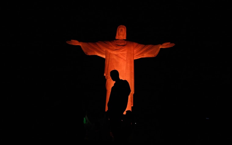 Image: Man stands in front of statue of Christ the Redeemer as it is lit up in orange to commemorate the Day against Human Trafficking and Missing Persons, celebrated by the Archdiocese of Rio de Janeiro