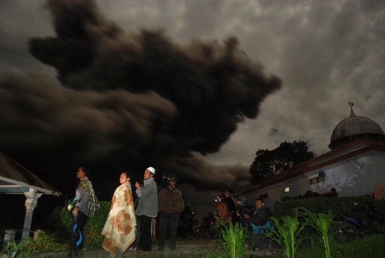 Image: Residents gather outside their houses in Karo district as dark giant ash clouds, seen in the background, rise from the crater of Mount Sinabung