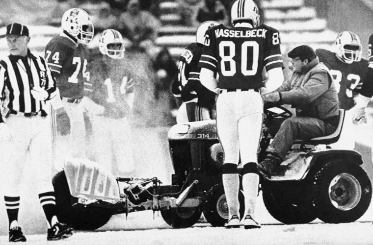 A worker at Schaefer Stadium in Foxboro drives a modified tractor along the sidelines to clear the snow away while members of the New England Patriots watch on Sunday, Dec. 12, 1982, during third quarter National Football League action with the Miami Dolphins. 