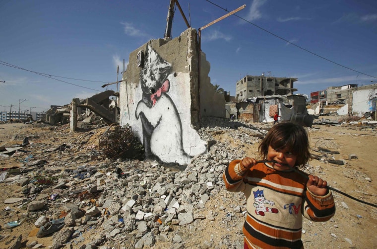 Image: Palestinian girl looks on as a mural of a playful-looking kitten, presumably painted by British street artist Banksy, is seen on the remains of a house wall, in Biet Hano