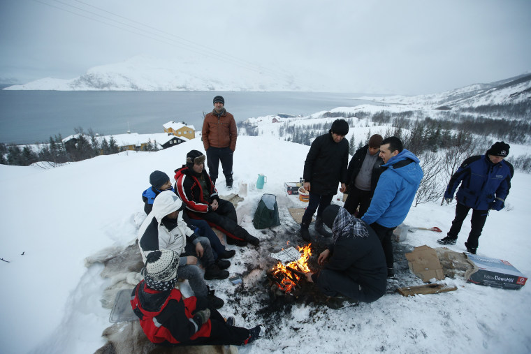 Image: Asylum seekers gather round a fire as they cook a meal overlooking the temporary Altnes camp refugee camp