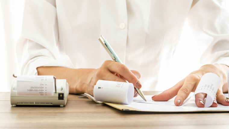 Front view of female bank employee in elegant white blouse writing something on receipts with documents and adding machine on her wooden desk.; Shutterstock ID 312203876; PO: today.com - mish